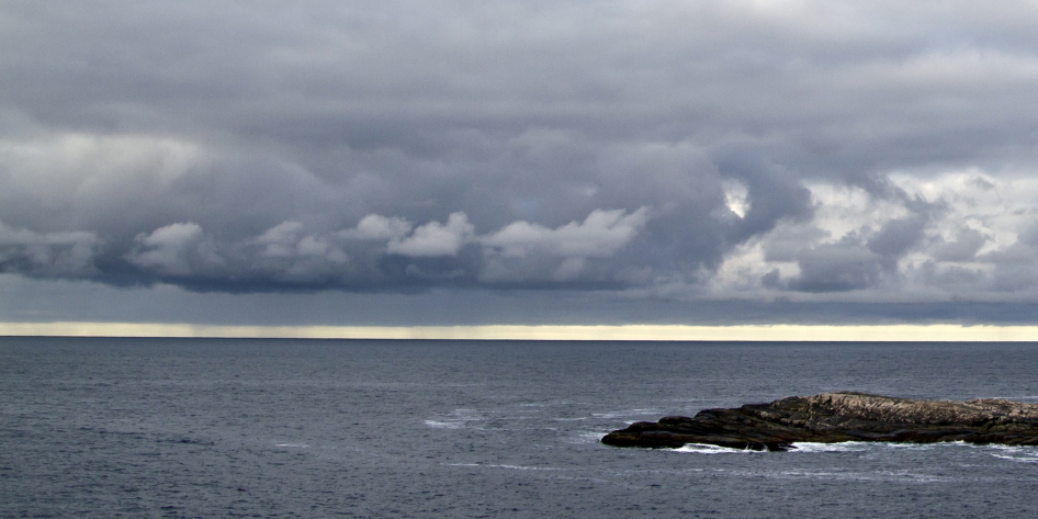 Mørke stormskyger som samler seg over et rolig hav ved en klippeformasjon.